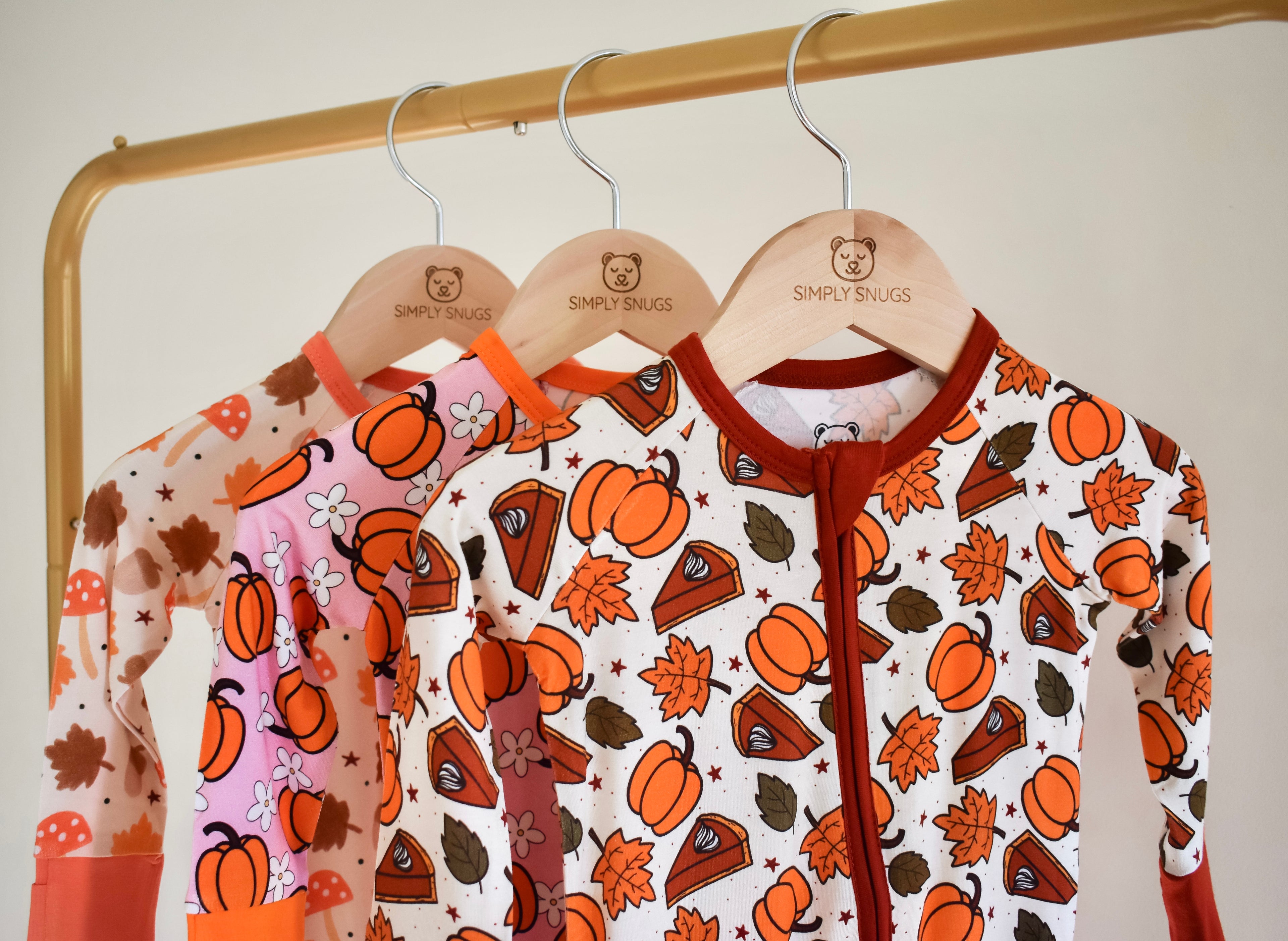 Three baby rompers with autumn-themed prints on wooden hangers.