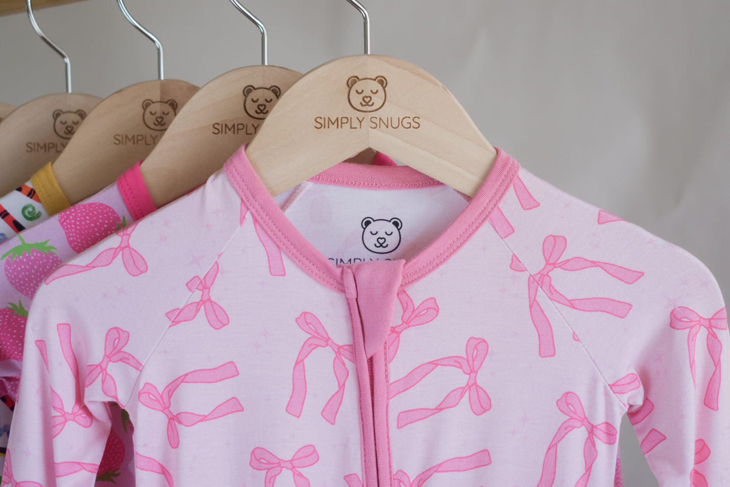Pink baby pajama with bow pattern on a hanger with 'Simply Snugs' branding.