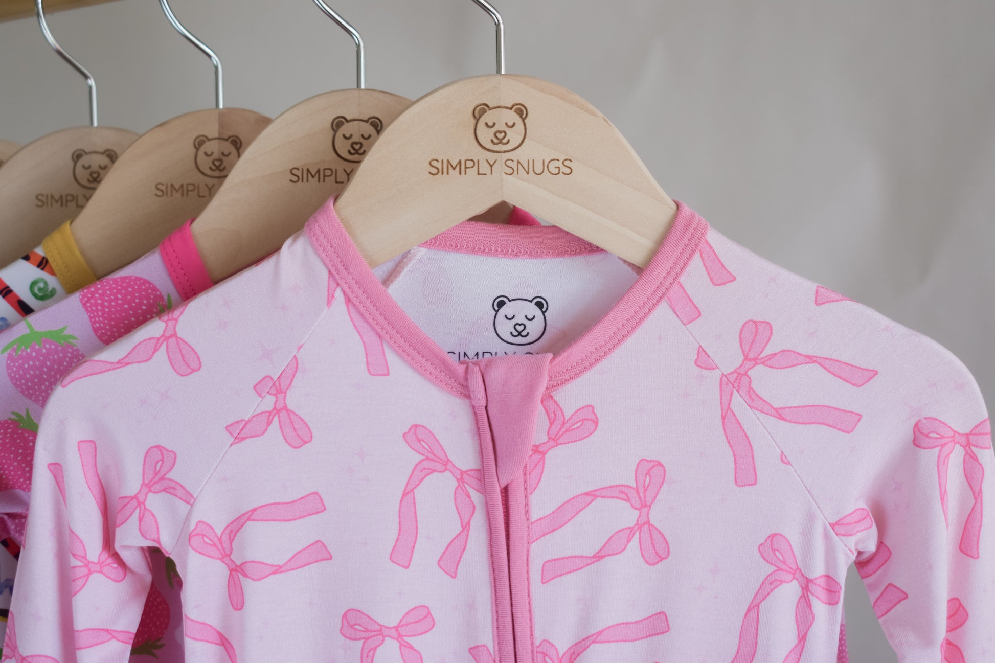 Pink baby pajama with bow pattern on a hanger with 'Simply Snugs' branding.