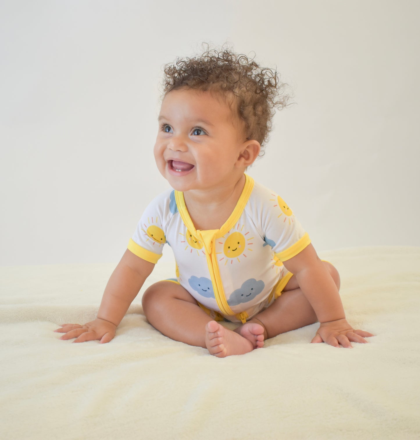 Happy baby sitting on a white blanket wearing a yellow and white bamboo pajama with cloud and sun patterns.