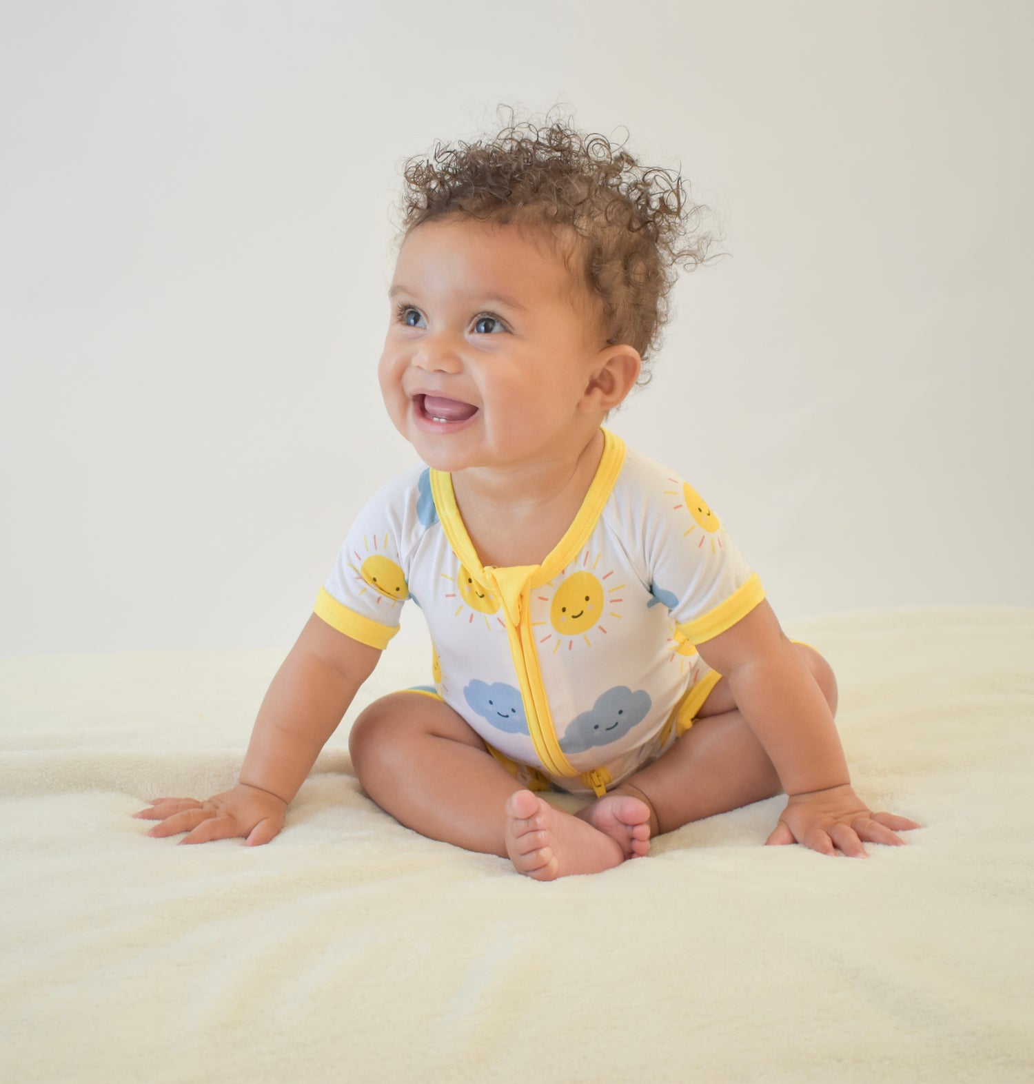 Happy baby sitting on a white blanket wearing a yellow and white bamboo pajama with cloud and sun patterns.